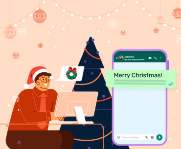 WhatsApp Christmas holiday messages: A brown man with short hair wearing a Santa hat sits in front of a computer and next to a Christmas tree. In a speech bubble coming out of his mouth, there is a wreath with a bow on top of it. The background is orange with various strings of lights and ornaments. On the right of the image, there is a smartphone opened to a WhatsApp chat with Katherine. The message from the sender in the chat says "Merry Christmas!", and the rest of the message is obscured from view.